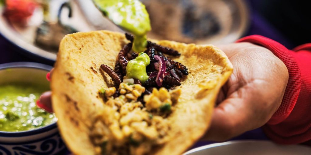 Hand holding a taco filled with guacamole, corn, onions, and fried mezcal worms, photo by Marco/Adobe Stock
