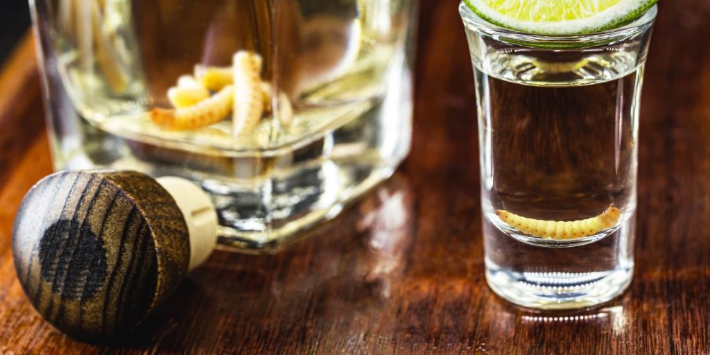 Mezcal shot glass with a gusano and lime wedge, next to a mezcal bottle also containing worms, photo by RHJ/Adobe Stock