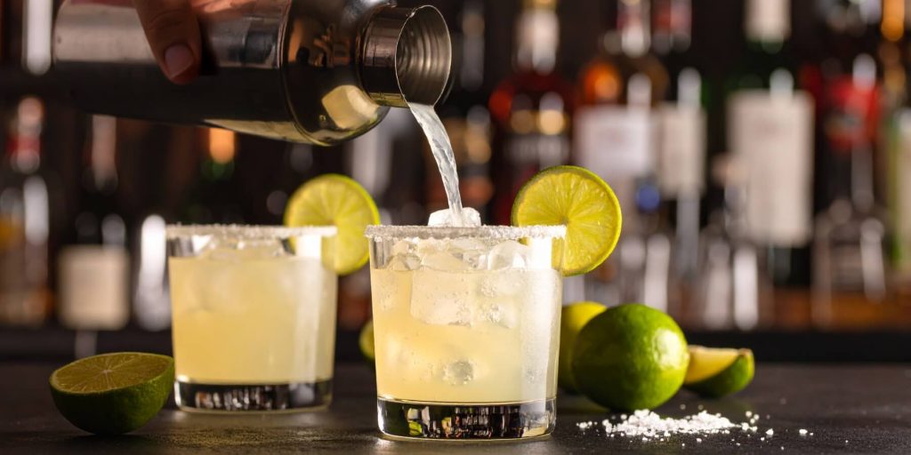 Close-up of a hand pouring a margarita into a salt-rimmed rocks glass, modern bar in the background