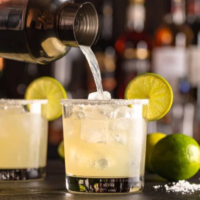 Close-up of a hand pouring a margarita into a salt-rimmed rocks glass, modern bar in the background