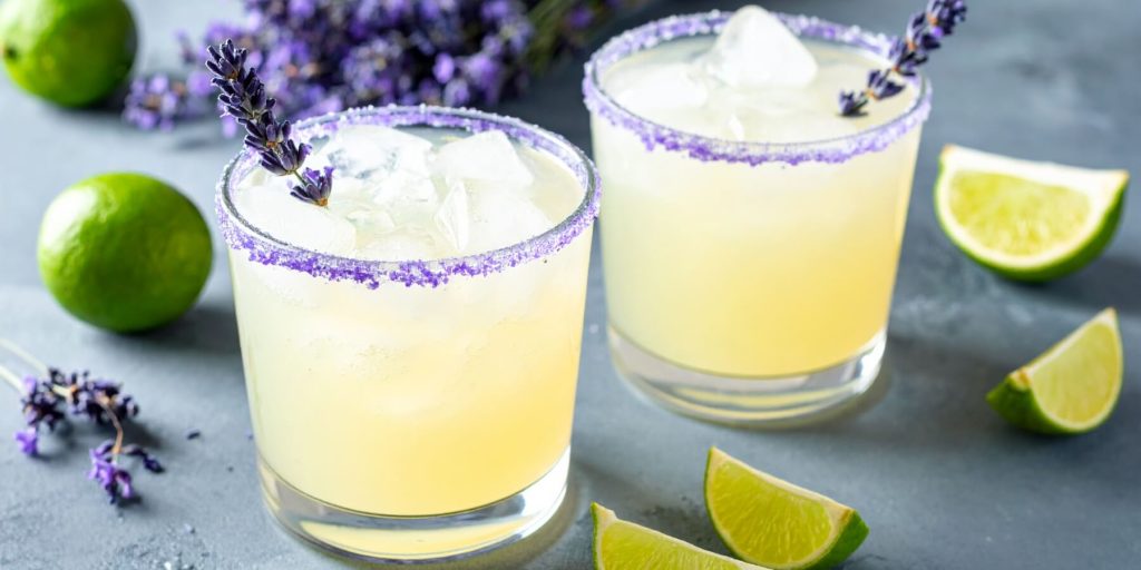 Two Lavender Margaritas with purple sugar rims and lavender sprigs, surrounded by lime wedges and fresh lavender.