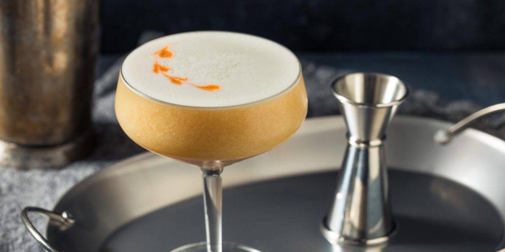 Beretta’s Rattlesnake cocktail in a coupe glass with a creamy foam and bitters garnish, accompanied by maple leaves on a rustic surface. Image by Brent Hofacker on Adobe Stock.