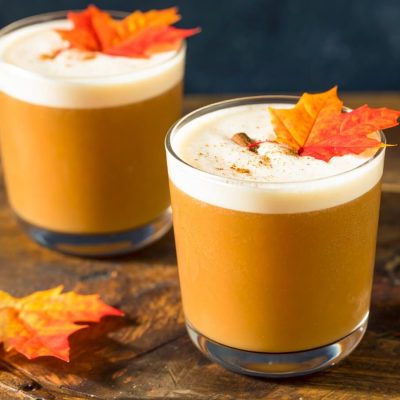 Two fall-themed cocktails with frothy tops in short glasses, garnished with cinnamon and maple leaves, set on a wooden surface.