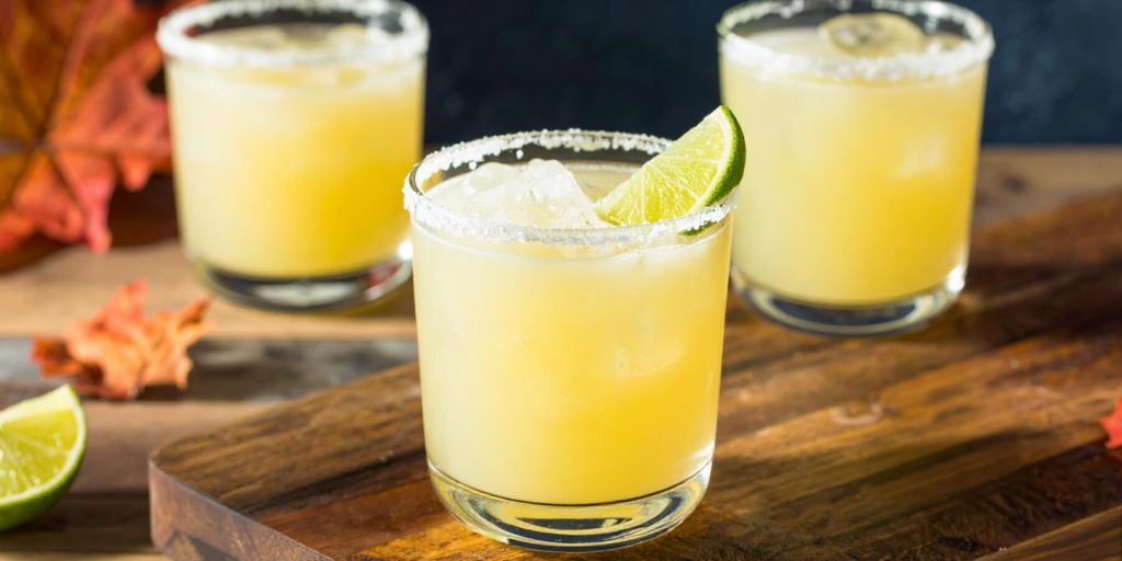 Three Maple Margarita cocktails with salted rims, garnished with a lime wedge each, on a wooden table in a home setting