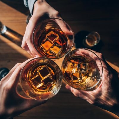 Three glasses of bourbon with ice being clinked over a table