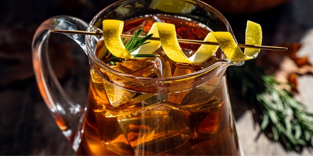 A close-up of a glass pitcher containing Maple Whiskey Sour Punch, a golden-brown Thanksgiving batch cocktail with large ice cubes and garnished with a dramatic lemon peel twist and a sprig of rosemary.