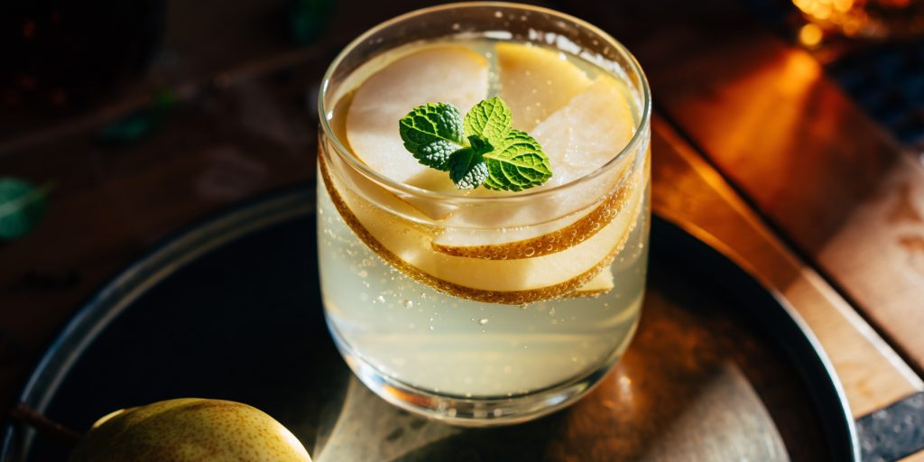 A sparkling Thanksgiving batch cocktail, Pear + Ginger Prosecco Punch, served in a short glass filled with pear slices and topped with a fresh mint sprig. The golden drink catches warm light on a dark wood tray.