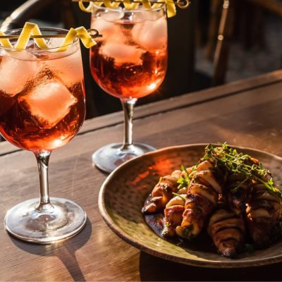 Two pink-hued Vincha Rosada cocktails in stemmed glasses garnished with curled lemon peels, set on a dark wooden table alongside a plate of bacon-wrapped appetizers. The golden-hour light enhances the warm tones of this Argentinian cocktail moment.