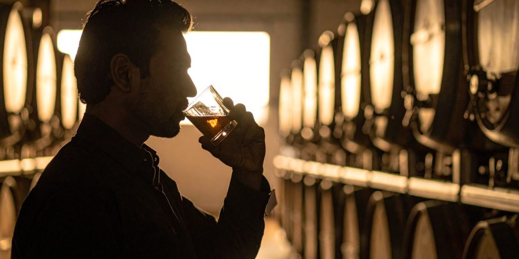 A side shot of a man taking a sip of bourbon out of a glass in a rickhouse