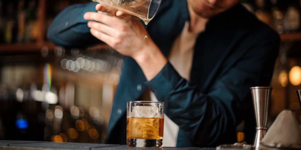 A bartender pouring a Tequila Old Fashioned out of a mixing jug into a rocks glass with ice.
