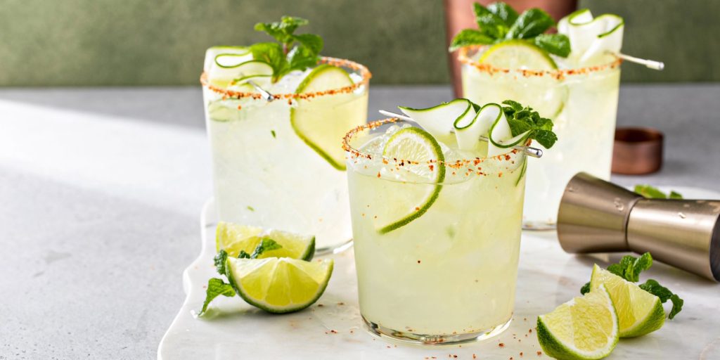 Three Margarita cocktails garnished with limes and cucumber ribbons on a marble surface.