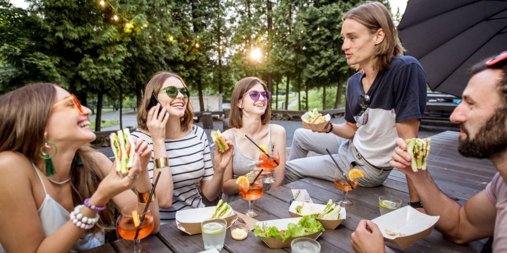 A group of friends sharing a picnic and Aperol Spritz cocktails on a park bench.