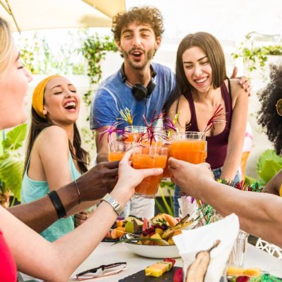 A group of friends toasting with cocktails outside around a picnic table with snacks