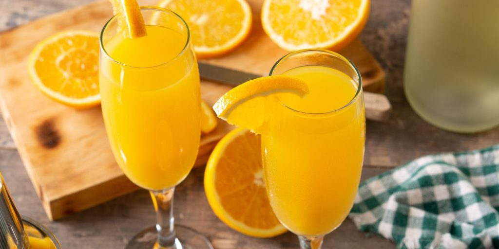 Close-up top view of two Mimosas garnished with fresh orange wedges, surrounded by more sliced oranges and assorted decor items including a chequered cloth